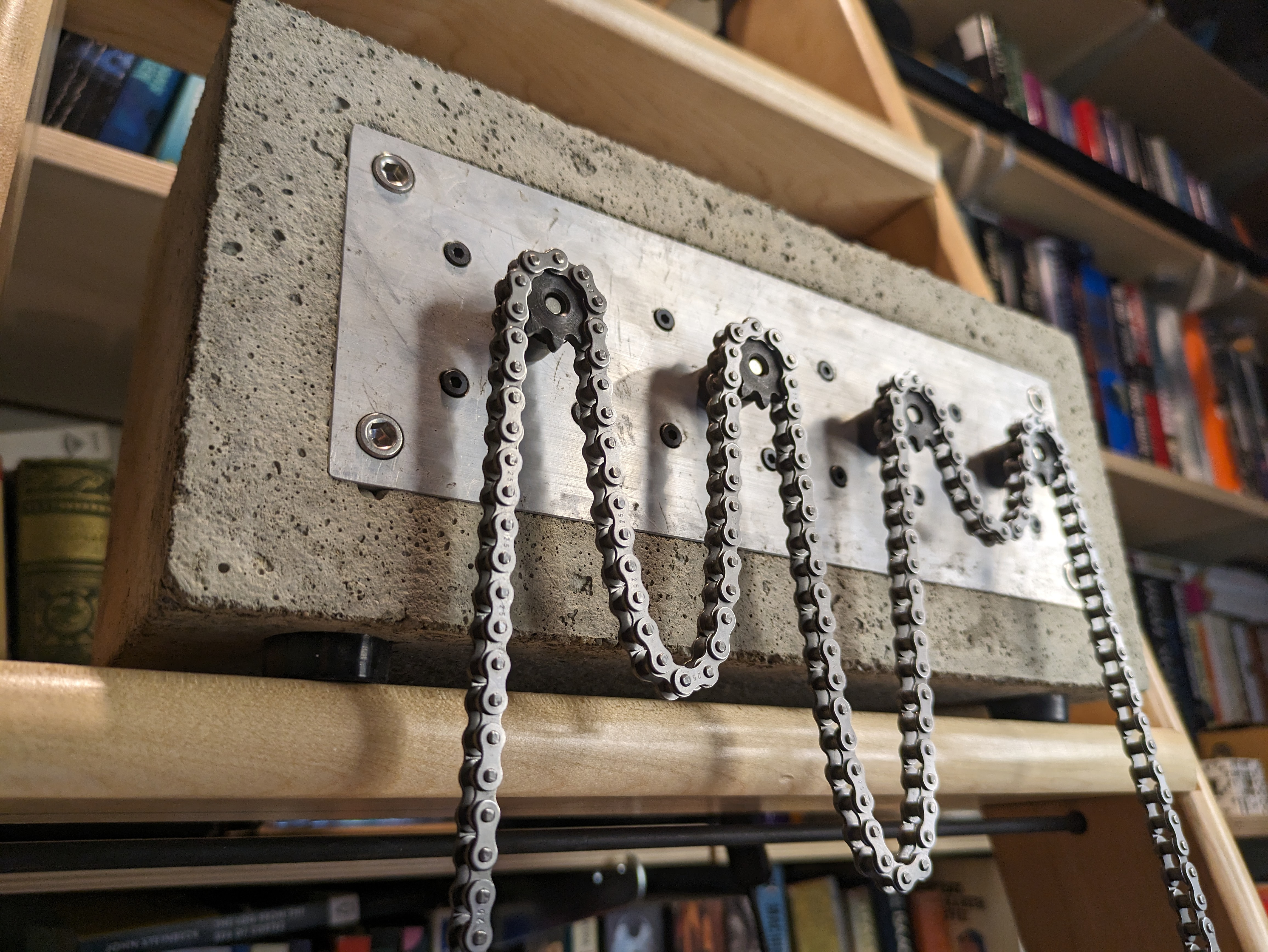 A concrete block sits on a library ladder. Embedded in the front face of the block is an aluminum plate, through which 4 small sprockets project. Over these a length of roller chain is draped, forming loops of various lengths between them.